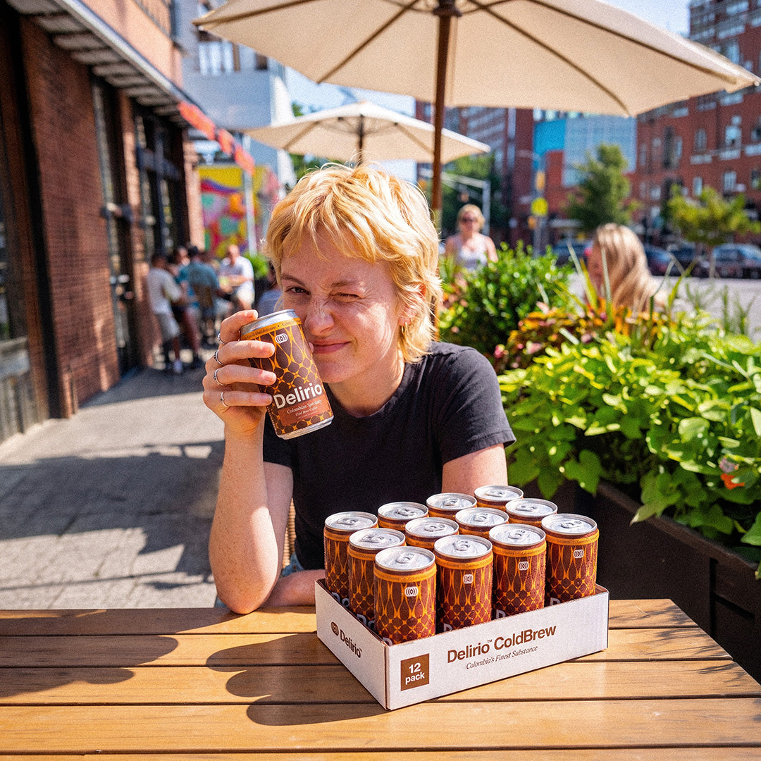 A woman sitting at an outdoor patio table holds a can of Delirio COLD BREW while a large box of Delirio COLD BREW cans sits on the table in front of her. The setting is a sunny urban street with brick buildings, green plants, and patio umbrellas.